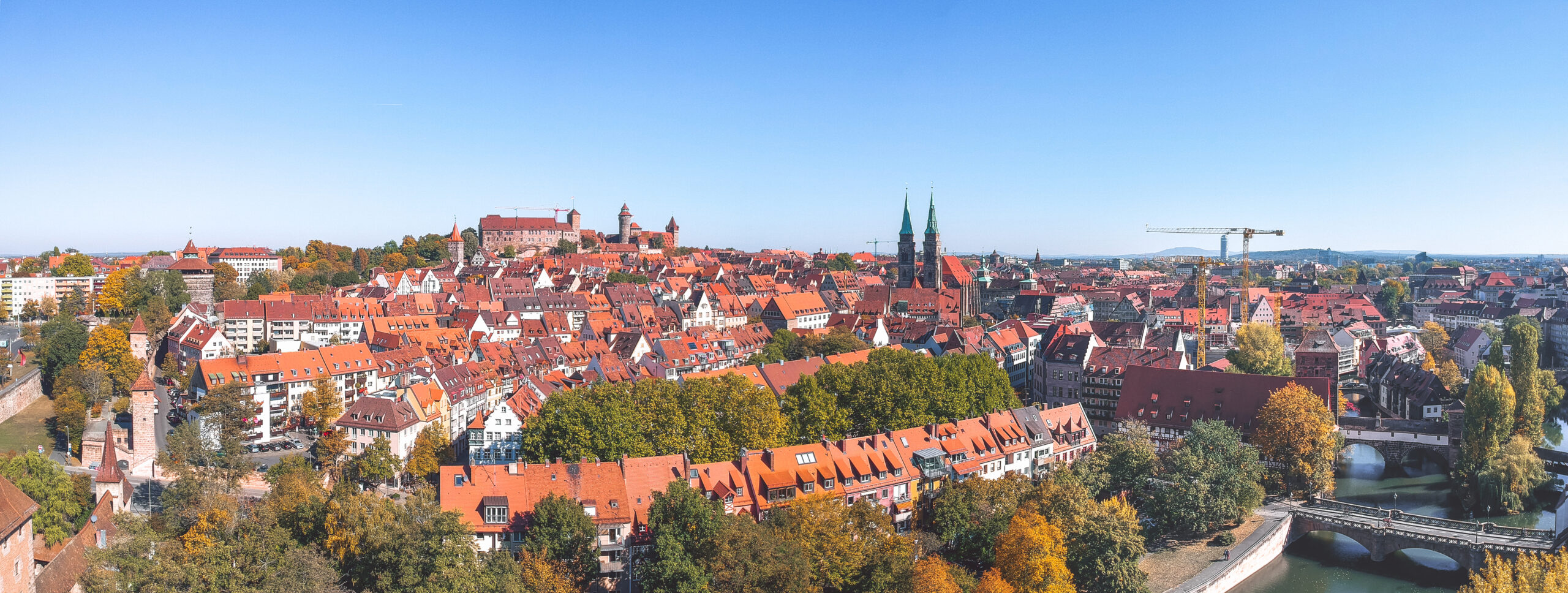 Historische europäische Stadt mit roten Dächern, Kirchtürmen, Bäumen und Fluss unter blauem Himmel in der Totalen.