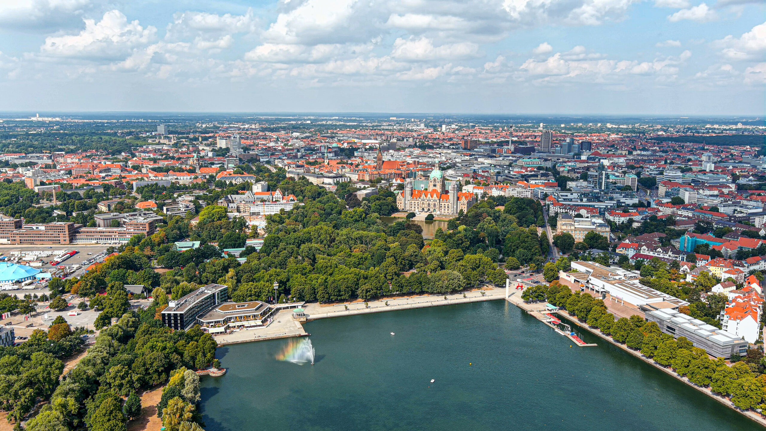 Stadt mit großem See, grünem Park und historischen Gebäuden aus der Vogelperspektive bei teilweise bewölktem Himmel.
