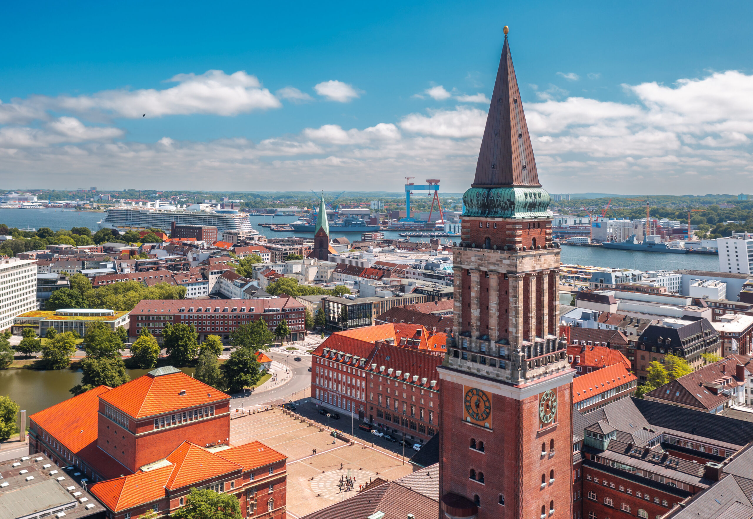 Backsteinuhrturm ragt über Stadt mit roten Dächern und Hafen, unter teils bewölktem Himmel.