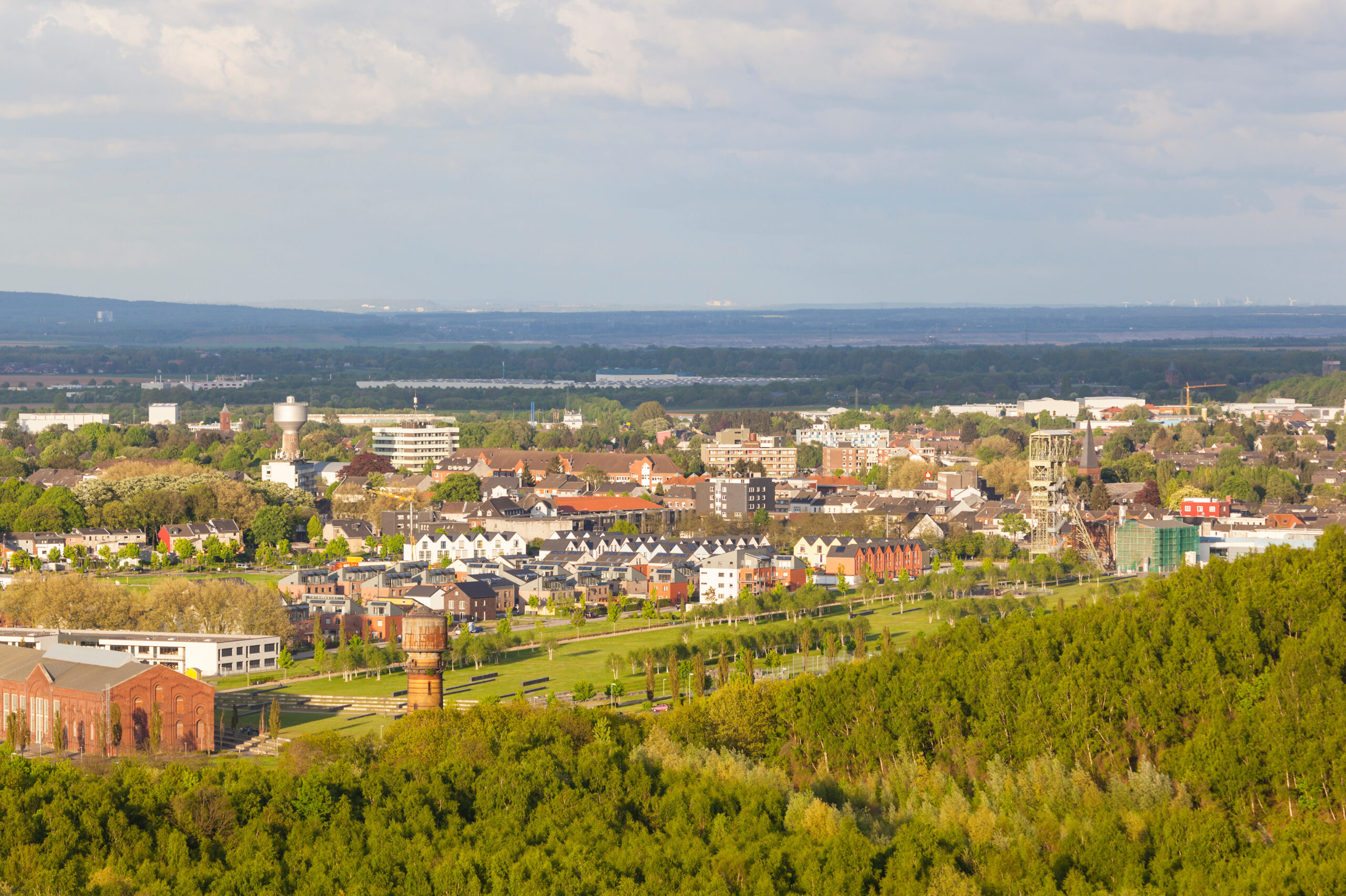 Luftaufnahme einer Stadt mit grünen Bäumen im Vordergrund und Gebäuden unter teils bewölktem Himmel.