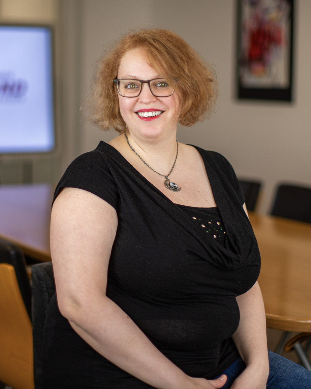 Frau mit Brille und schulterlangem Haar sitzt lächelnd in einem Konferenzraum mit Holztisch und Holzstühlen.