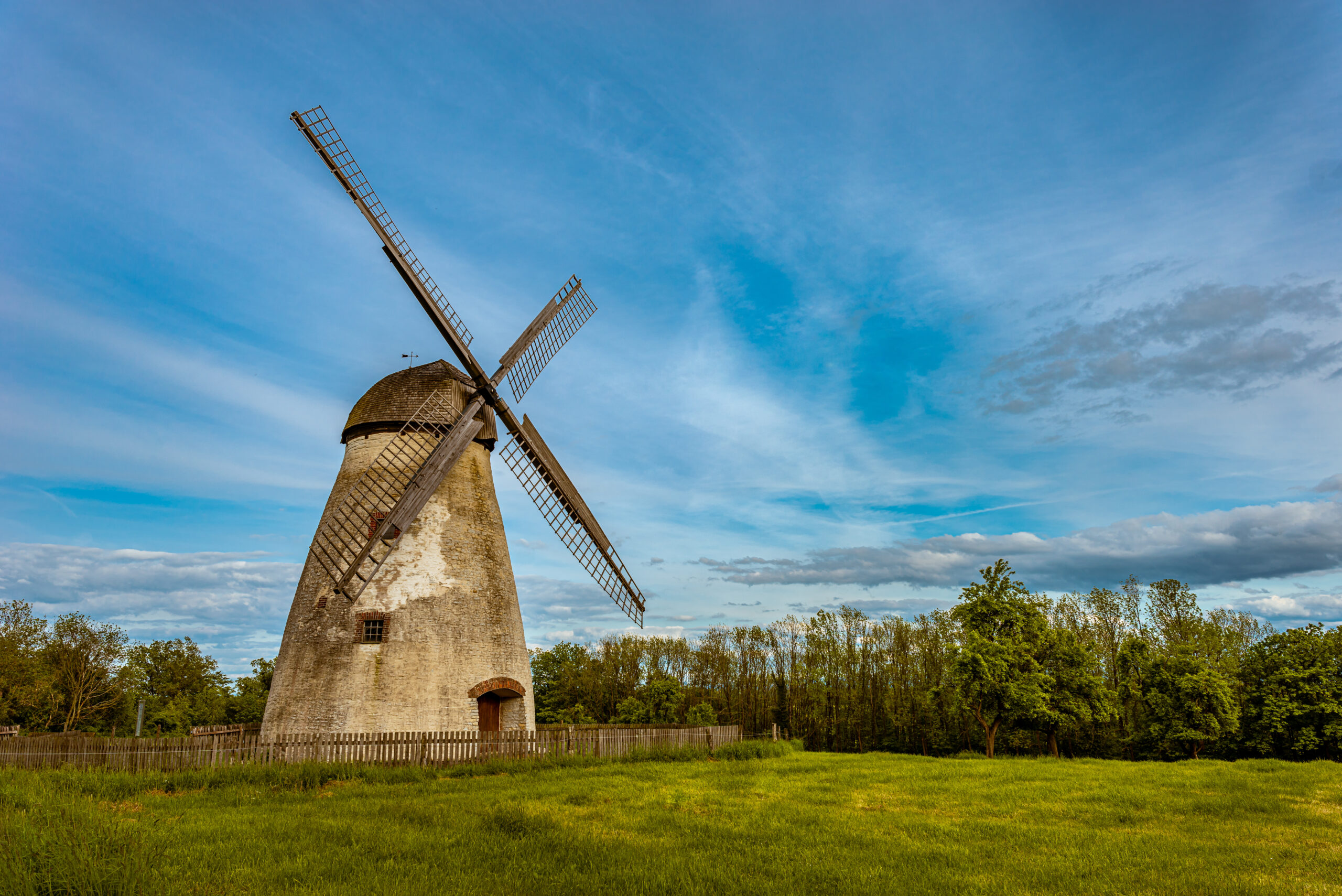Alte Windmühle steht auf grünem Feld unter blauem Himmel mit Wolken, umgeben von Bäumen.
