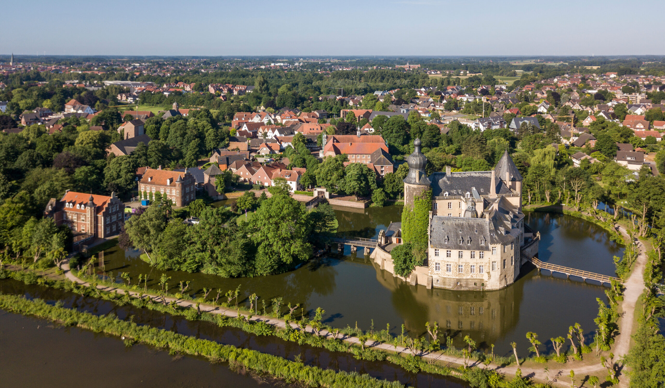 Luftaufnahme einer historischen Burg, umgeben von einem Wassergraben, mit Dorf und Grünflächen im Hintergrund.