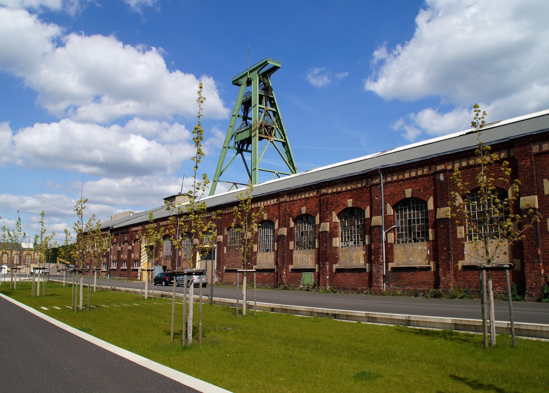 Backstein-Industriegebäude mit grünem Förderturm, junge Bäume an Straße, Himmel blau.