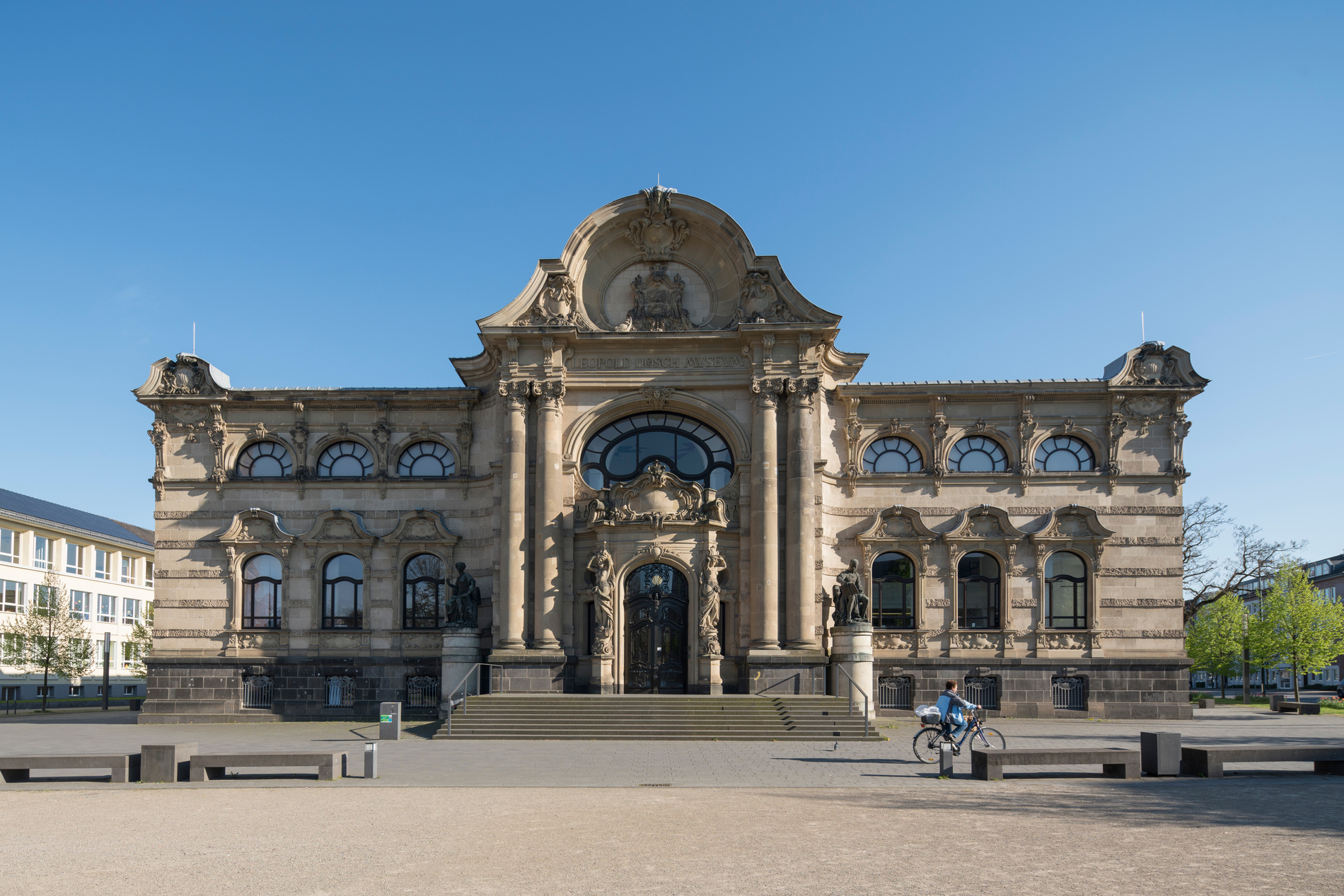 Radfahrer fährt an einem historischen Gebäude mit Rundbogenfensterstern und großem Eingang bei blauem Himmel vorbei.