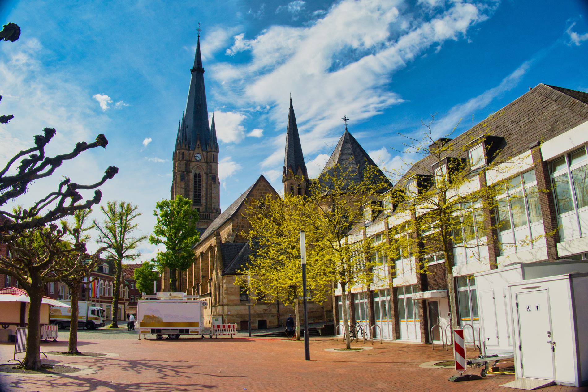Marktstände stehen auf sonnigem Stadtplatz mit Kirche, Laubbäumen und umliegenden Gebäuden unter blauem Himmel mit Wolken.