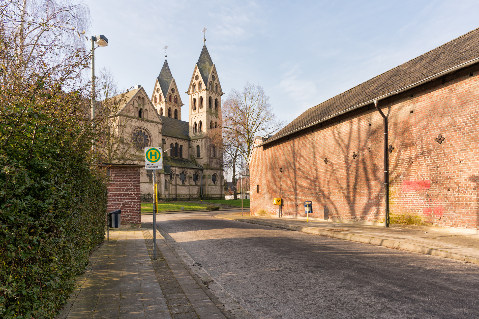 Backsteinkirche mit Doppelturm steht an ruhiger Straße neben Bushaltestelle bei sonnigem Wetter.