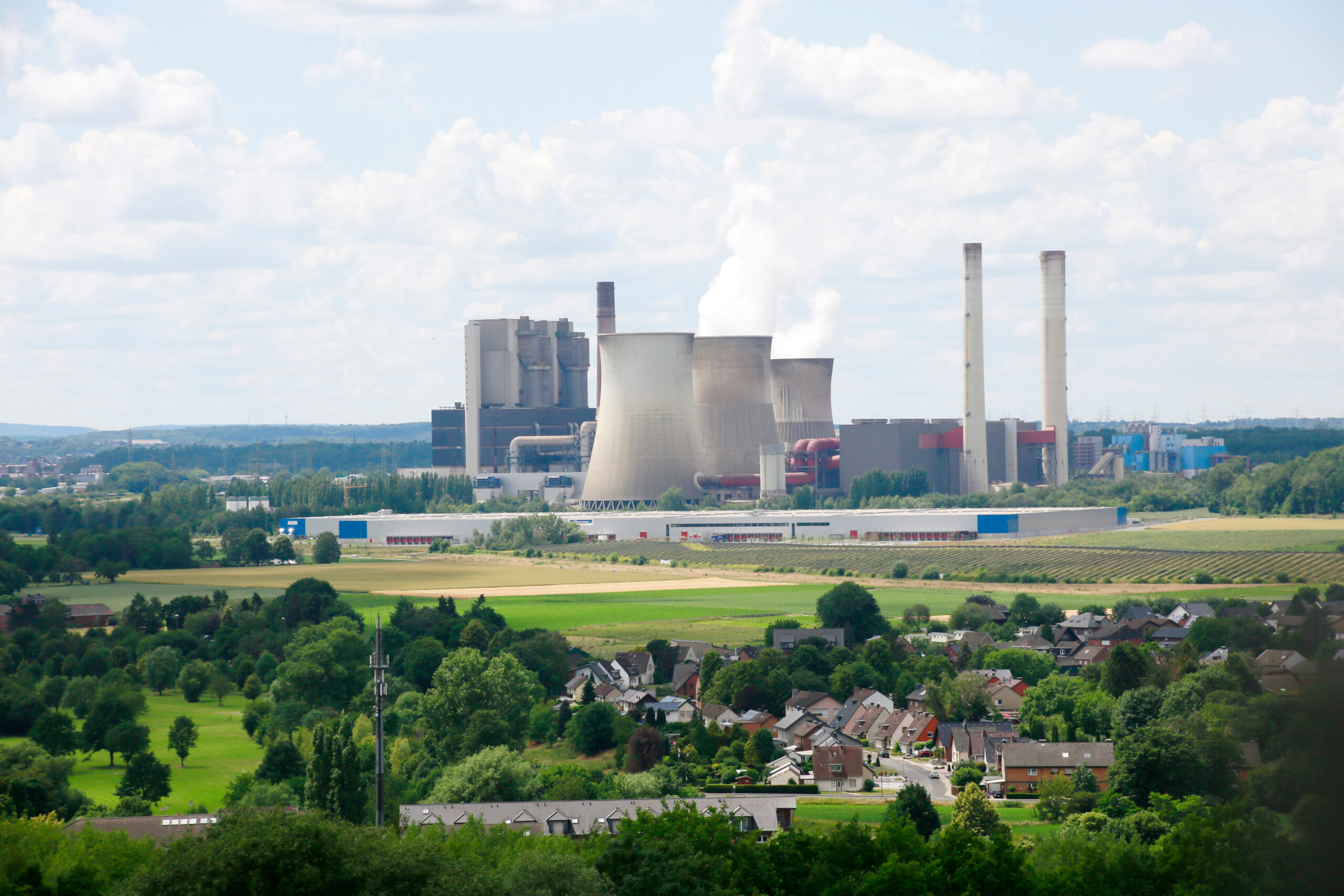 Kraftwerk mit Kühltürmen und Schornsteinen steht hinter grüner Landschaft mit Häusern und Bäumen.
