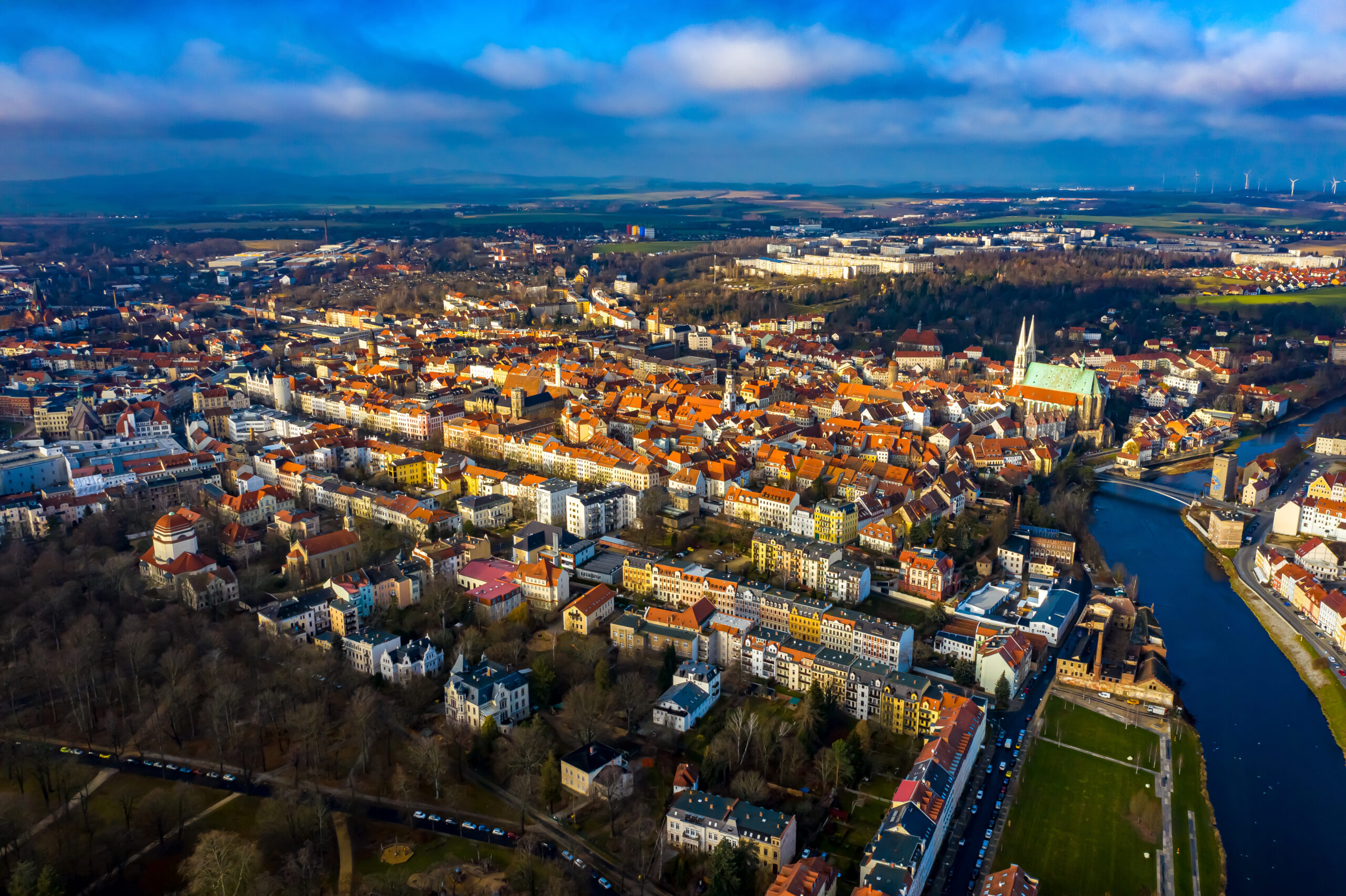 Luftaufnahme einer europäischen Stadt mit roten Dächern, Fluss und Grünflächen unter teils bewölktem Himmel.