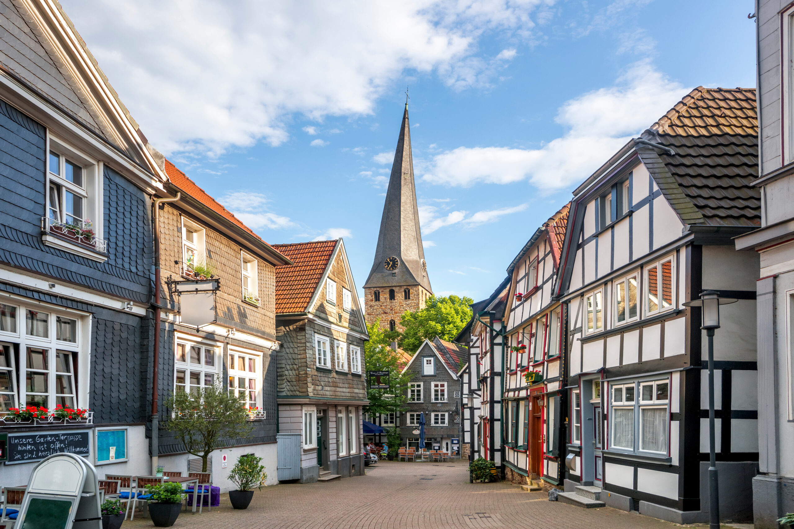 Kopfsteinpflasterstraße mit Fachwerkhäusern, im Hintergrund steht eine Kirche mit hohem Turm.