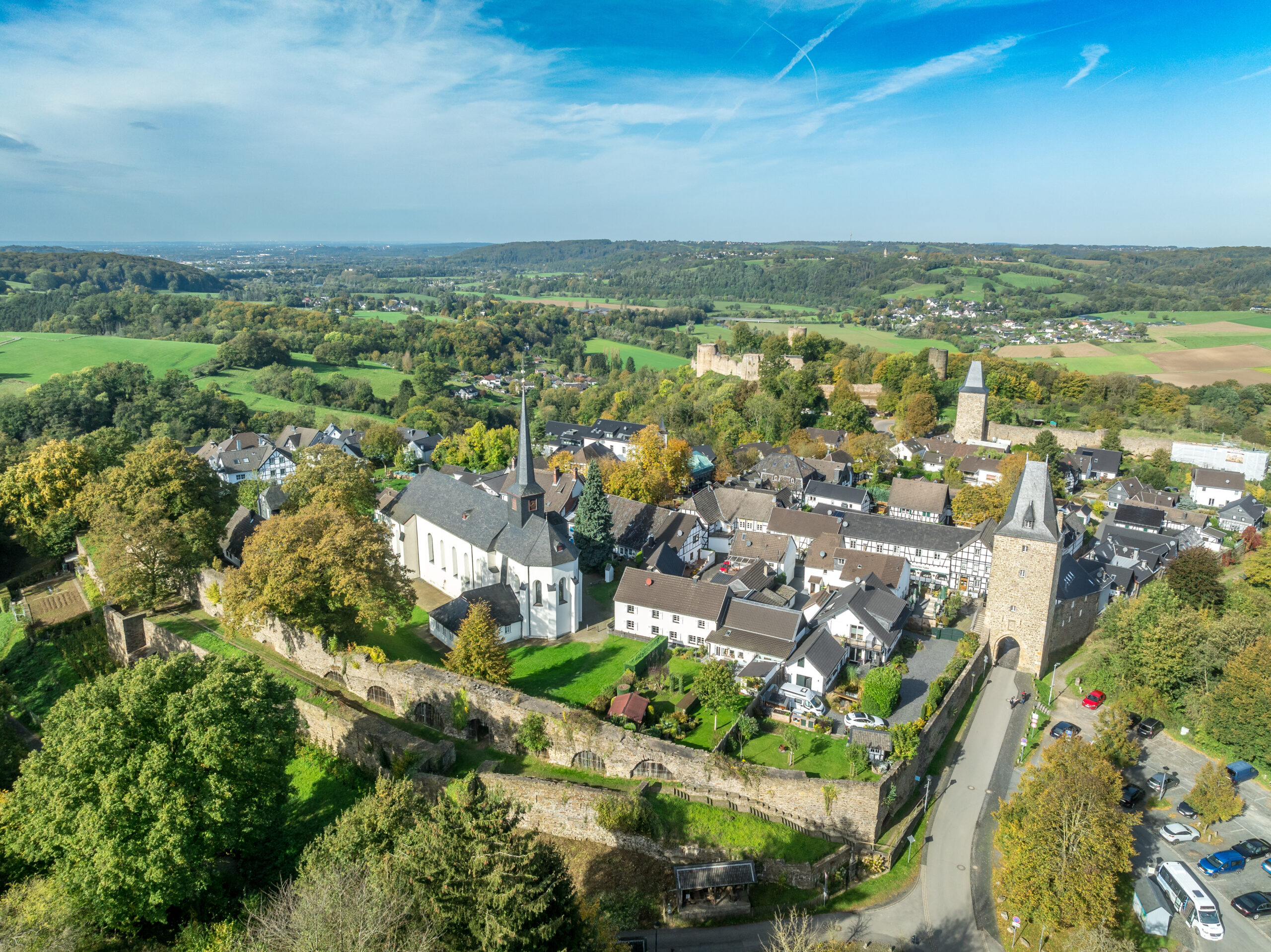 Luftaufnahme einer Kleinstadt mit Kirche, Steinturm und viel Grün bei blauem Himmel.
