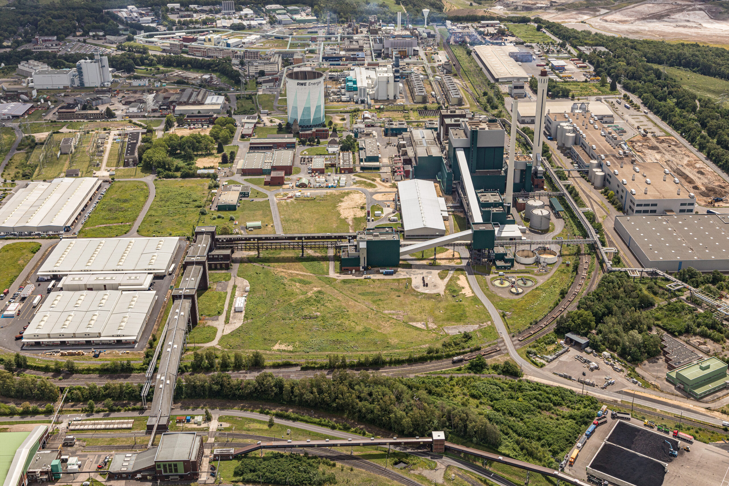 Luftaufnahme eines Industriegebiets mit Fabriken, Lagerhallen, Schornsteinen und angrenzendem Grün.