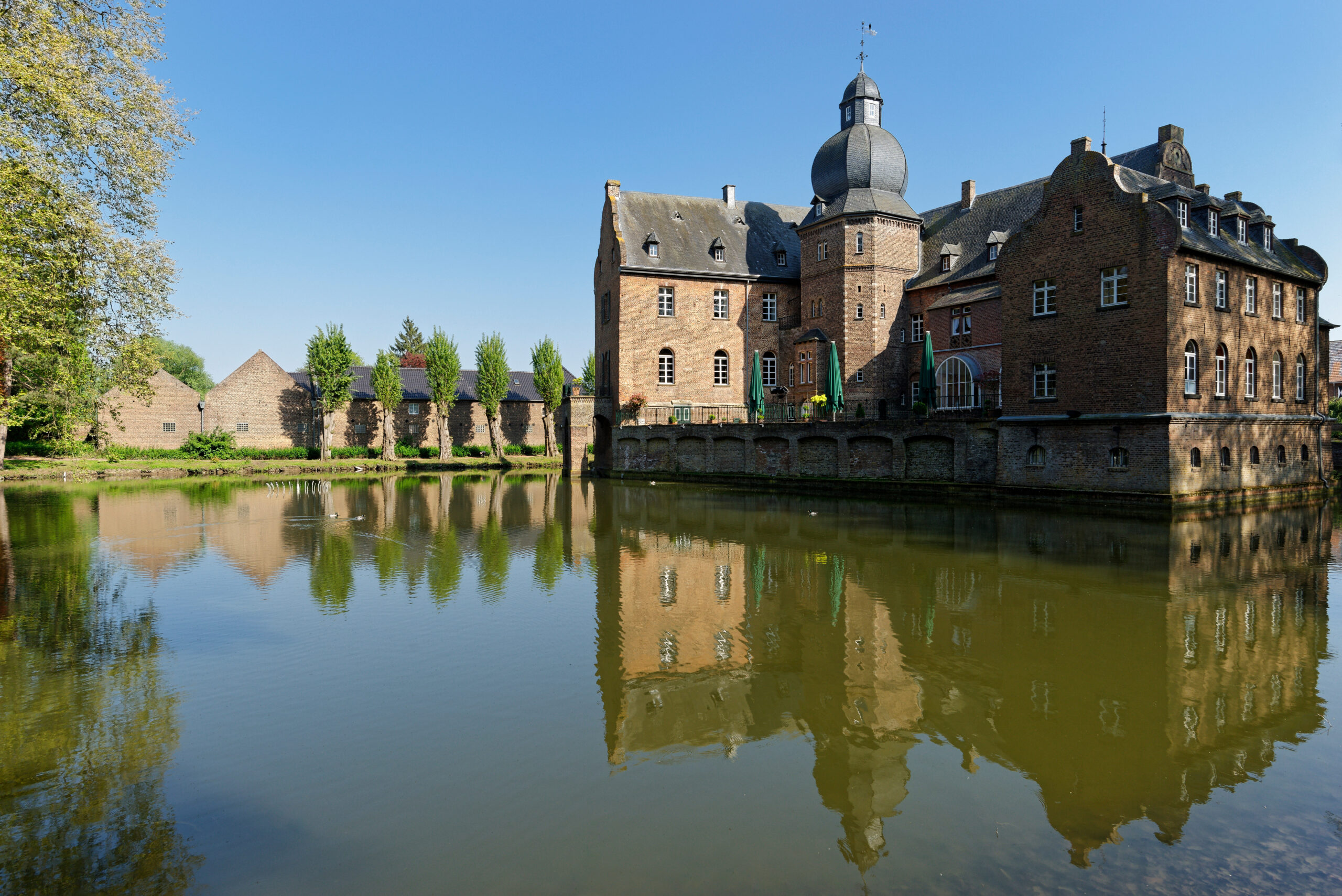 Backsteinburg mit Kuppelturm spiegelt sich in ruhigem Wassergraben, umgeben von Grün unter blauem Himmel.