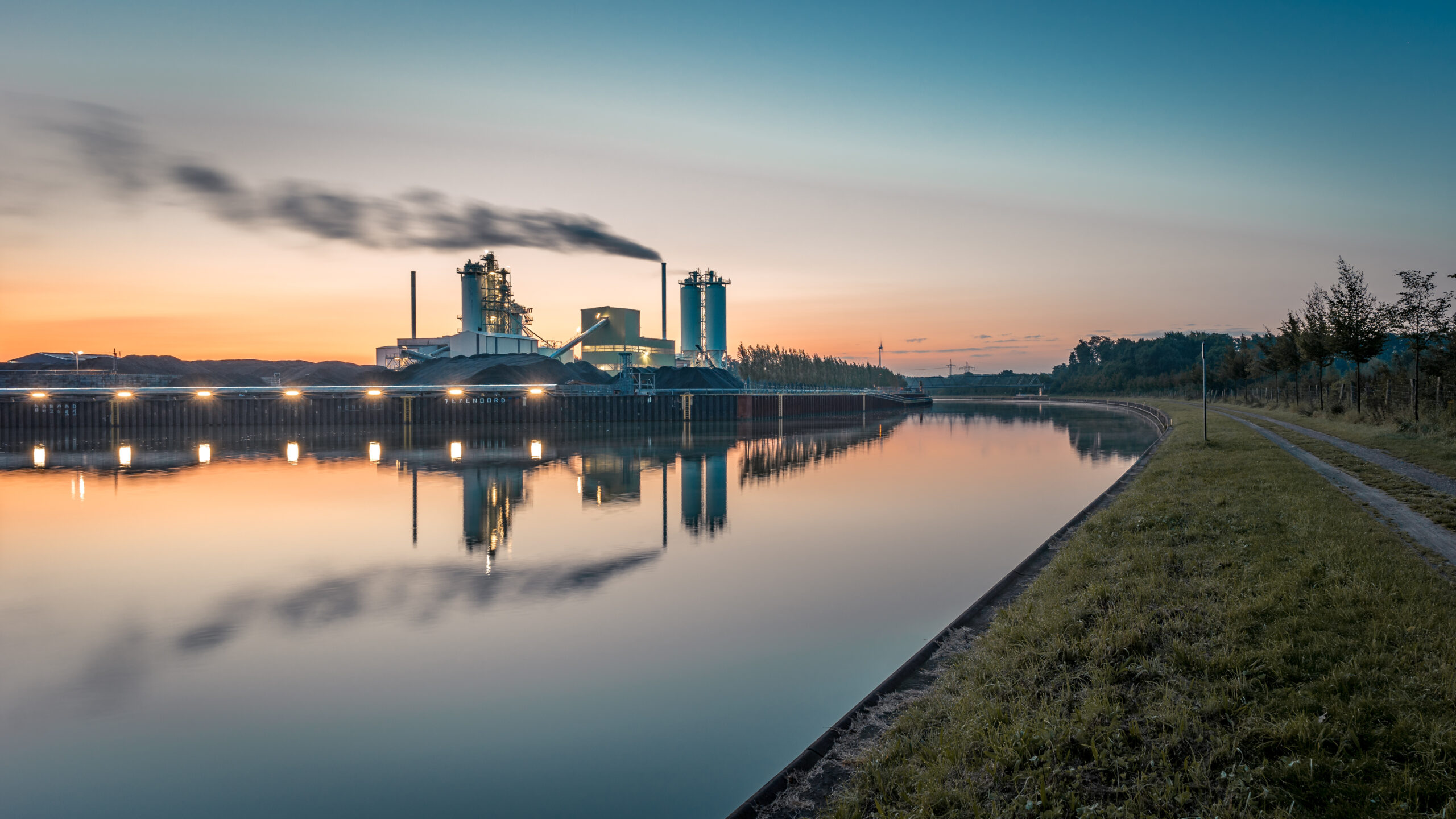 Fabrik stößt Rauch aus neben ruhigem Fluss bei Sonnenuntergang, Lichter spiegeln im Wasser, grasiger Weg daneben.