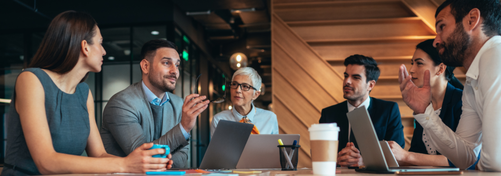 Sechs Personen im Business-Outfit besprechen eine Existenzgründung an einem Tisch mit Laptops im modernen Büro.
