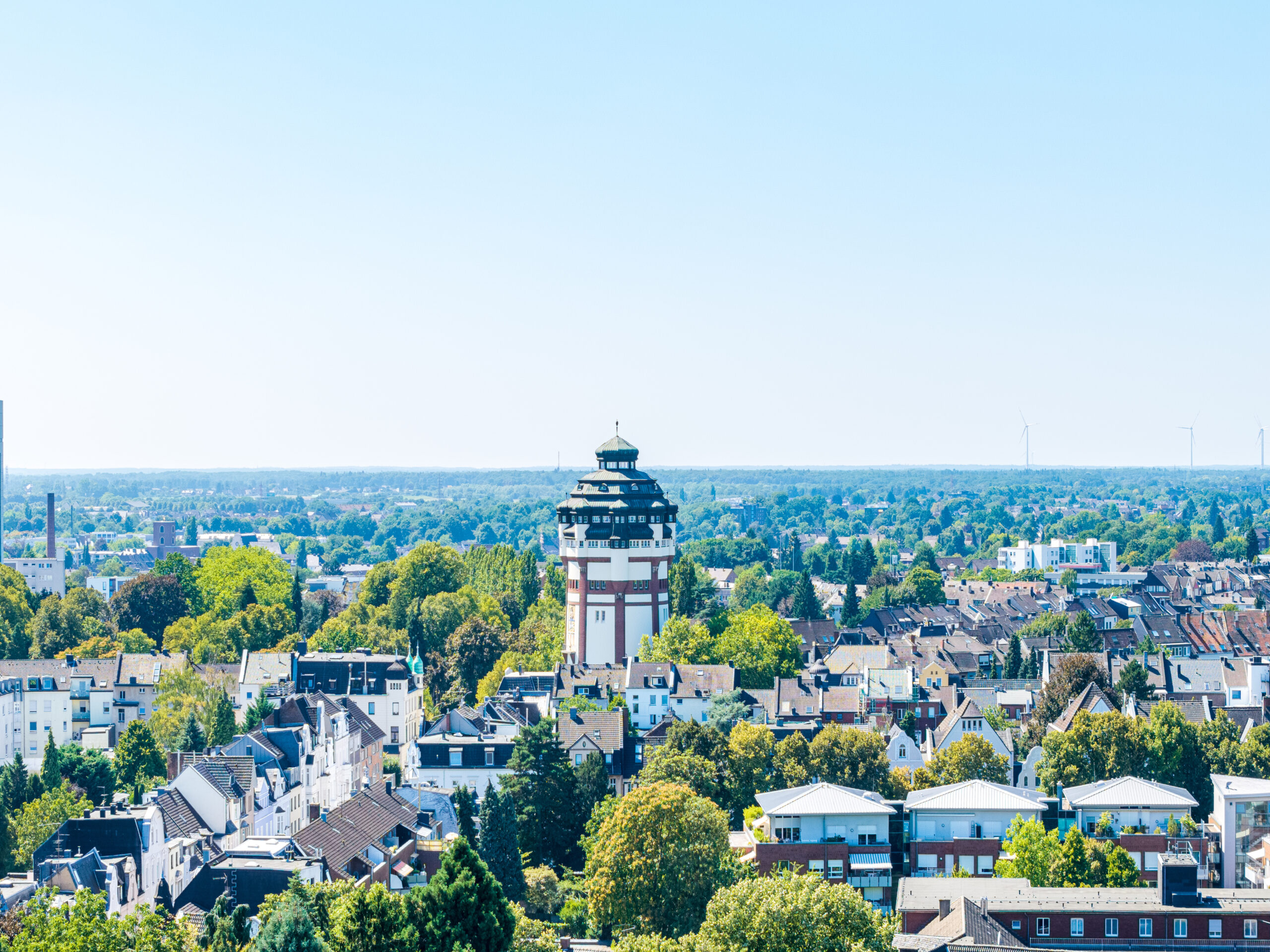 Luftaufnahme einer Stadt mit historischem Turm, umgeben von Bäumen und Wohnhäusern bei klarem Himmel.