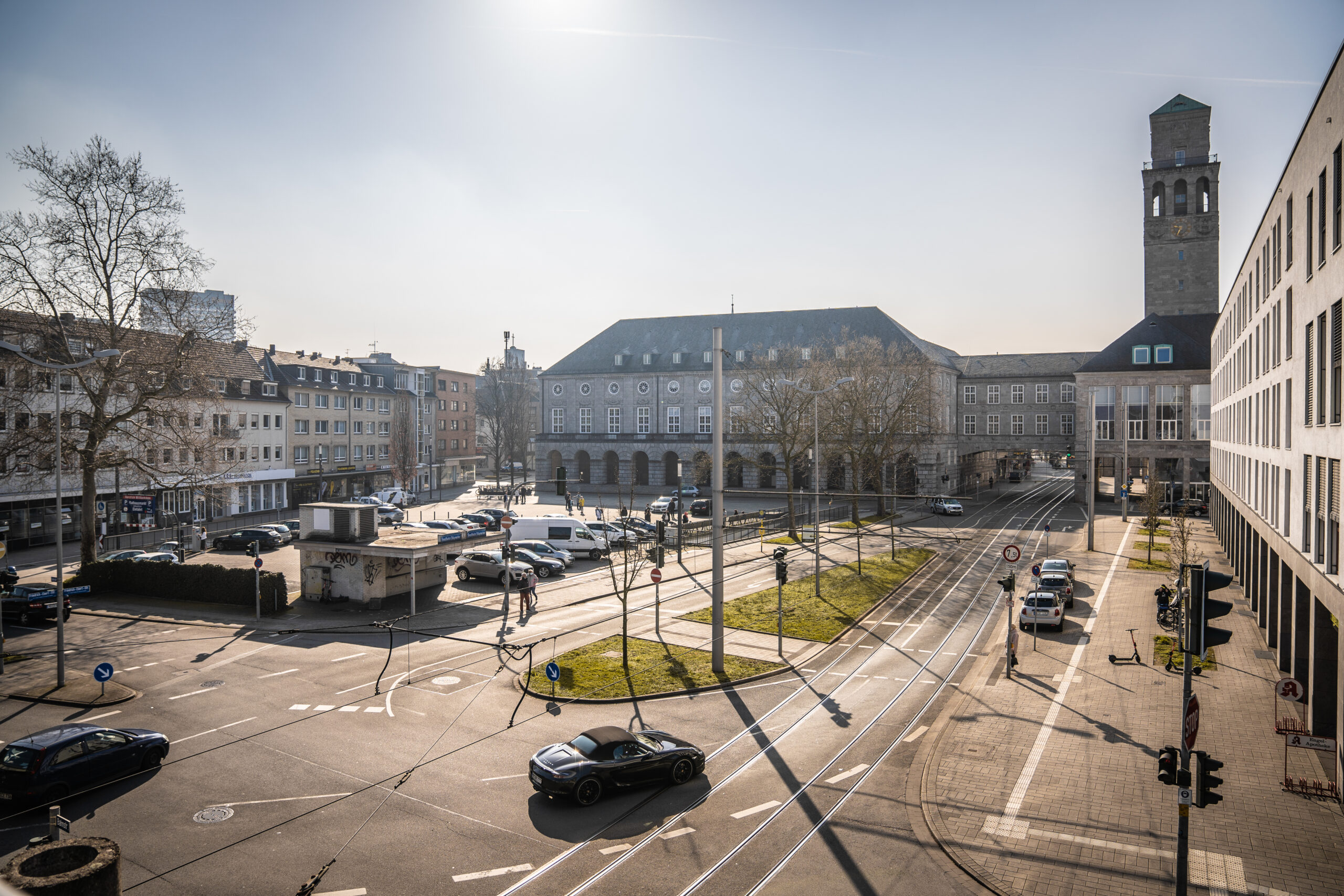Autos fahren bei Sonnenschein auf einen Stadtplatz mit historischen Gebäuden, Straßenbahnschienen und Uhrturm.