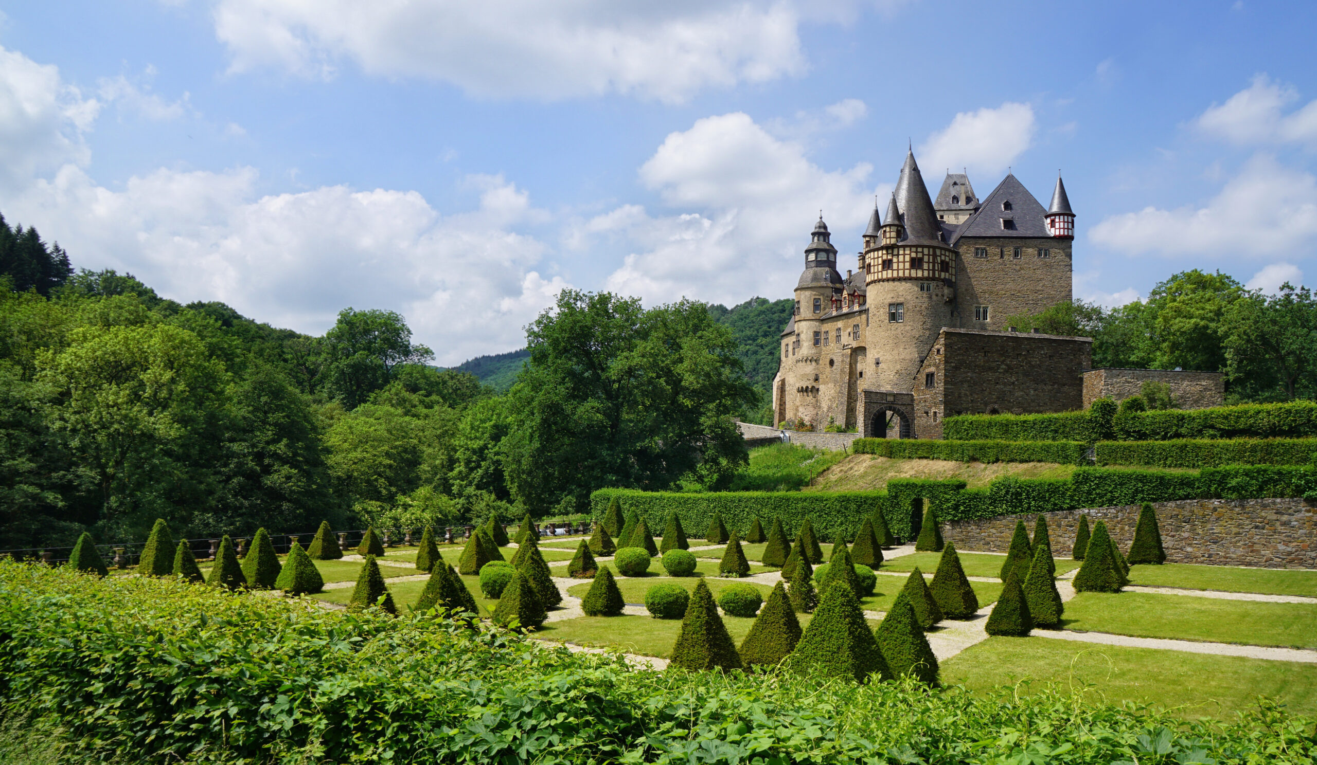 Steinburg mit Türmen steht über gepflegtem Garten mit Kegelschnitt-Sträuchern und viel Grün unter blauem Himmel.
