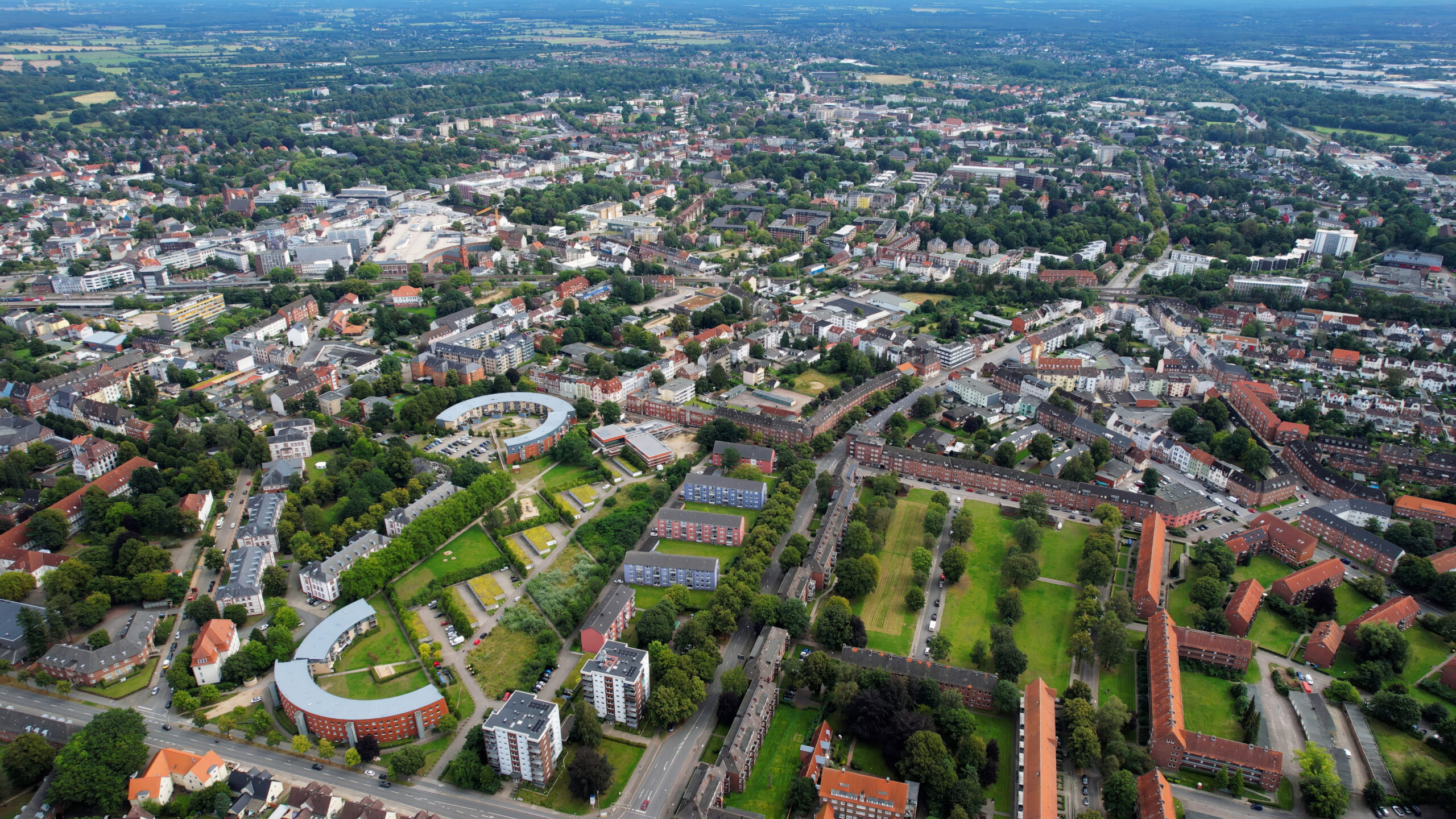 Luftaufnahme einer Stadt mit Wohngebäuden, Grünflächen und von Bäumen gesäumten Straßen unter blauem Himmel.