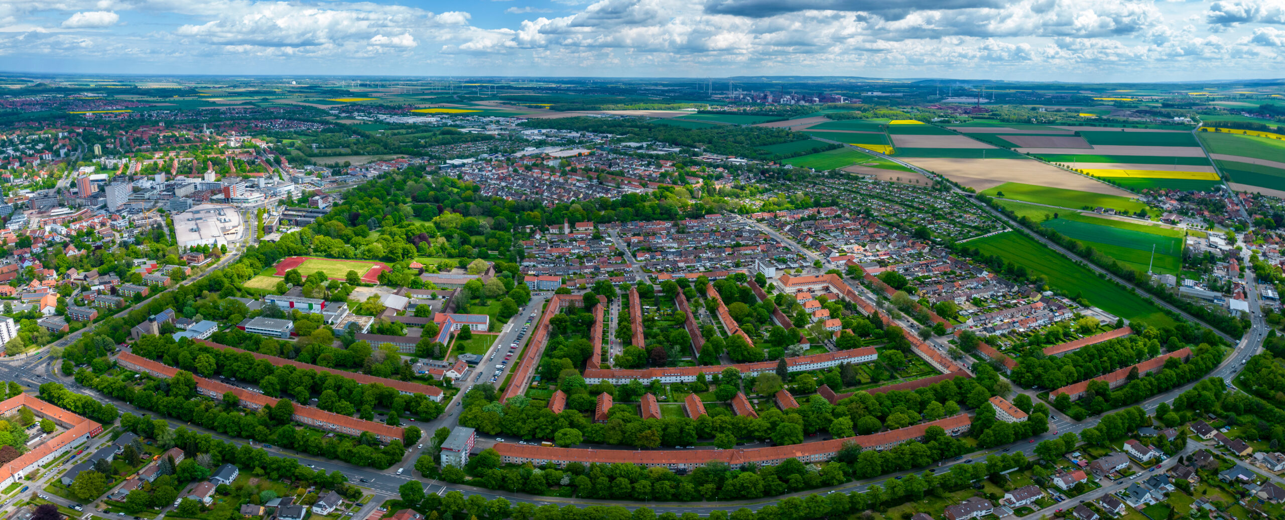 Luftaufnahme einer Ortschaft mit Häusern, grünen Bäumen, Feldern und bewölktem Himmel im Hintergrund.